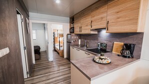 A compact kitchen with wooden cabinets, a countertop with a coffee maker, and a dining area visible through an open door. - Lake Serenity Premium Mobile Homes With Thermal Riviera Tickets (Brežice)