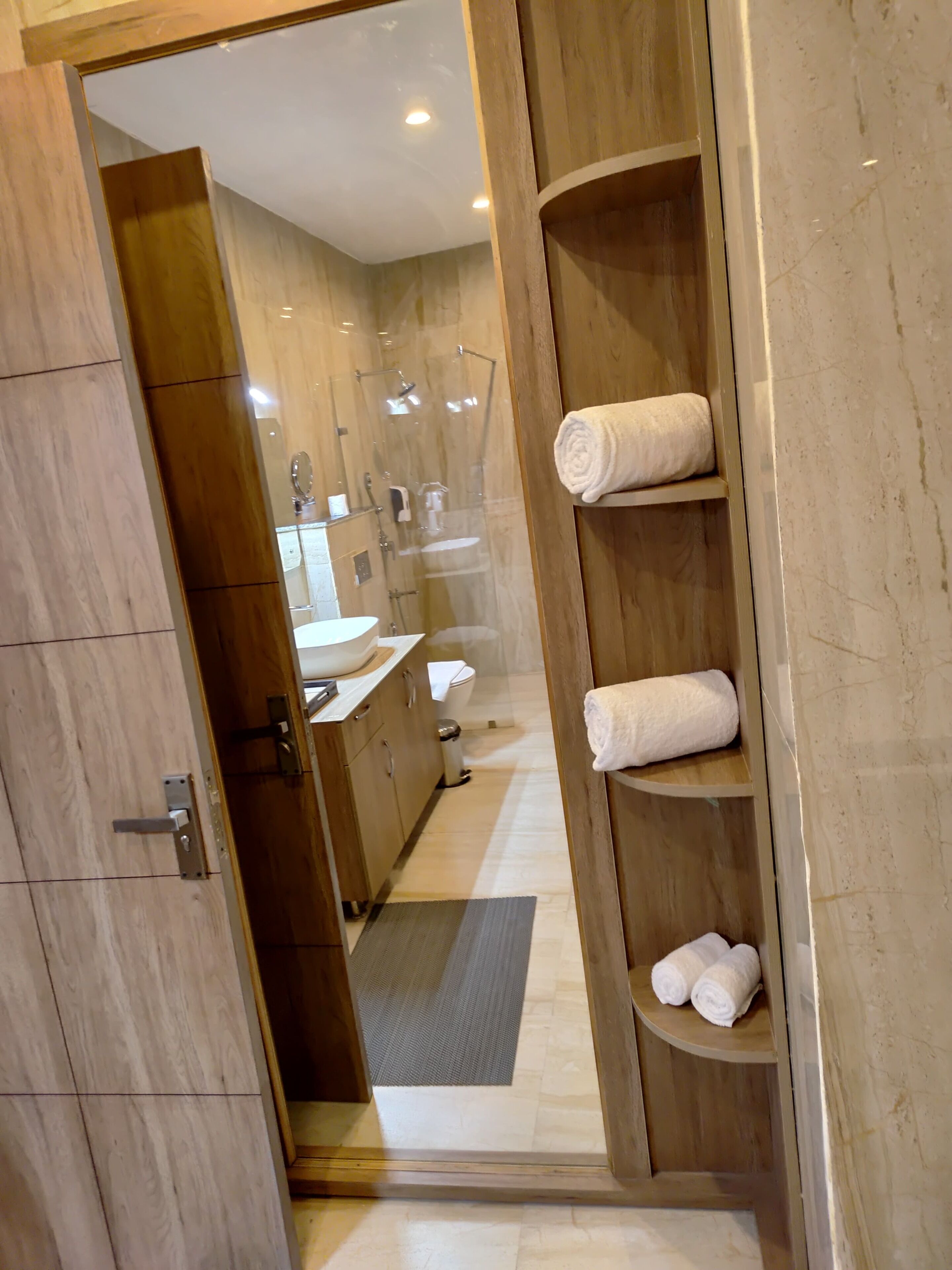 Une salle de bain moderne avec une douche à parois de verre, des étagères en bois où sont rangées les serviettes et un lavabo bien visible.