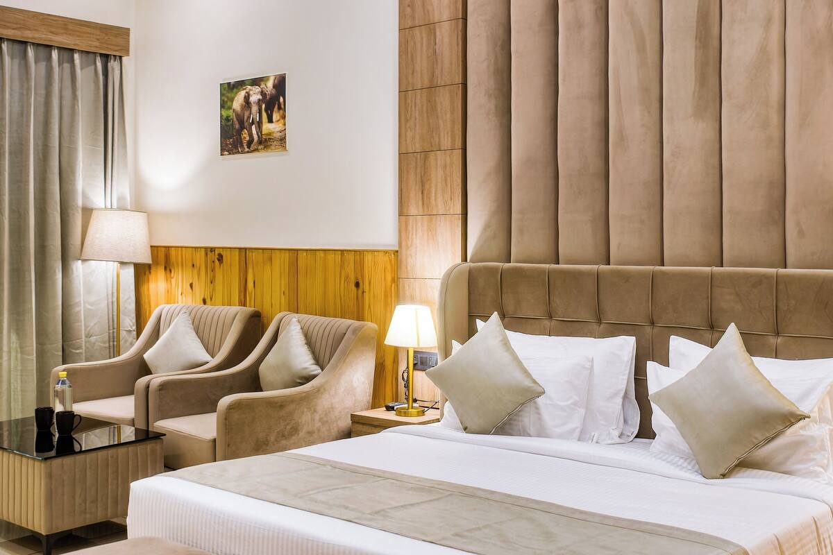 A hotel room with a large bed, two armchairs, a bench, a wooden panel wall, and a ceiling fan.