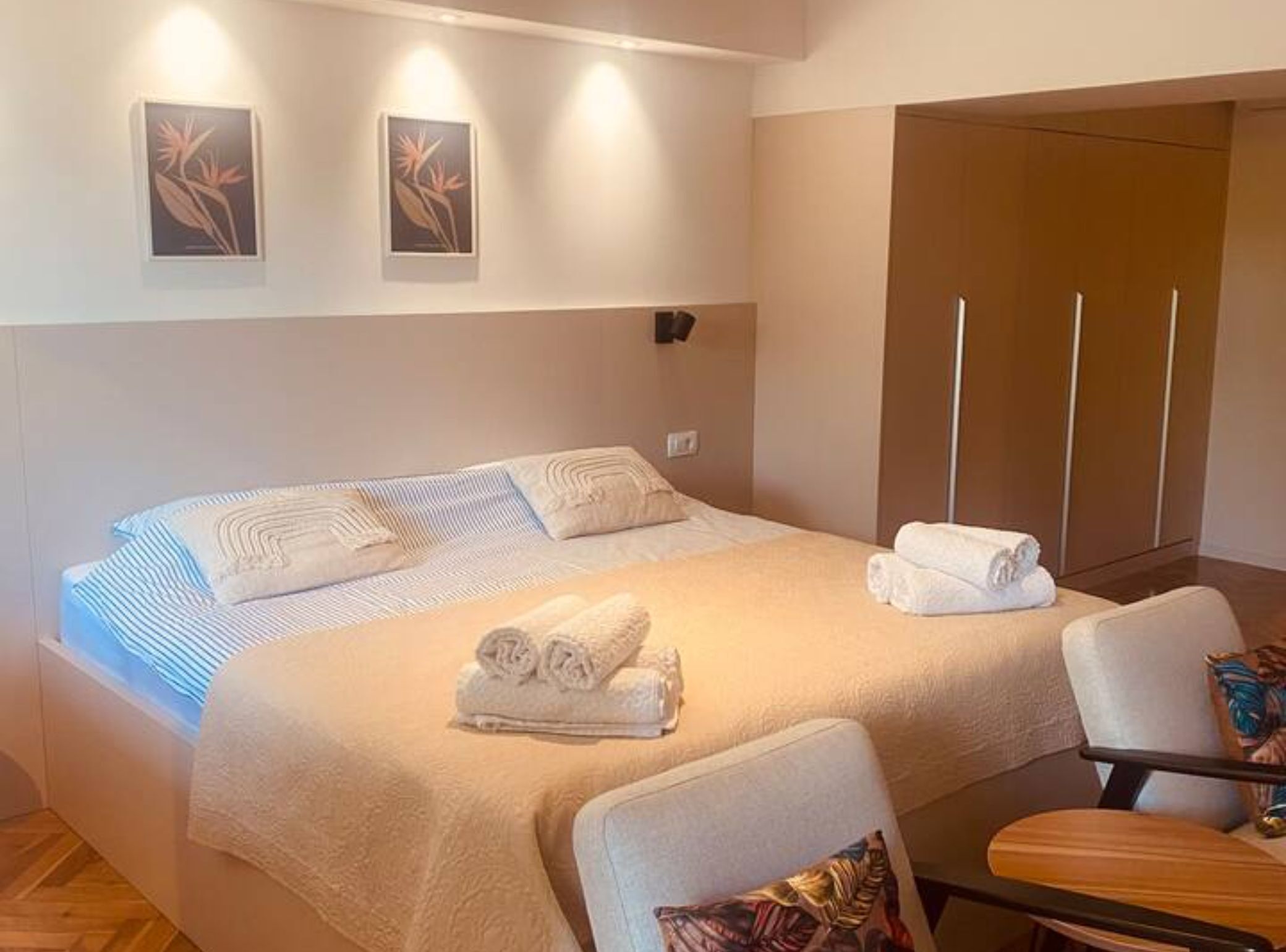 Un lit bien fait, recouvert d’une couvre-lit à rayures, avec deux oreillers et deux serviettes enroulées posées sur le lit. Deux appliques murales et deux tableaux encadrés ornent le mur. On aperçoit, au premier plan, une chaise en bois avec un coussin.