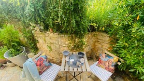 Une terrasse extérieure avec une table en pierre et deux chaises, entourée d’une végétation luxuriante et d’un mur en pierre.