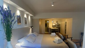 Une chambre d’hôtel moderne avec un grand lit, un coin salon comprenant un fauteuil et une petite table, et une kitchenette à l’arrière-plan.