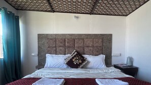 A hotel room with a large bed, a headboard, a nightstand, a kettle, and a window with curtains. - Crane Valley Cottage (Leh)