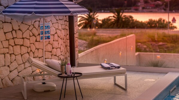Un espace détente avec un mur en pierre, un parasol bleu et blanc à rayures, une petite table avec une bouteille et un verre, et une vue sur une étendue d’eau au coucher du soleil.