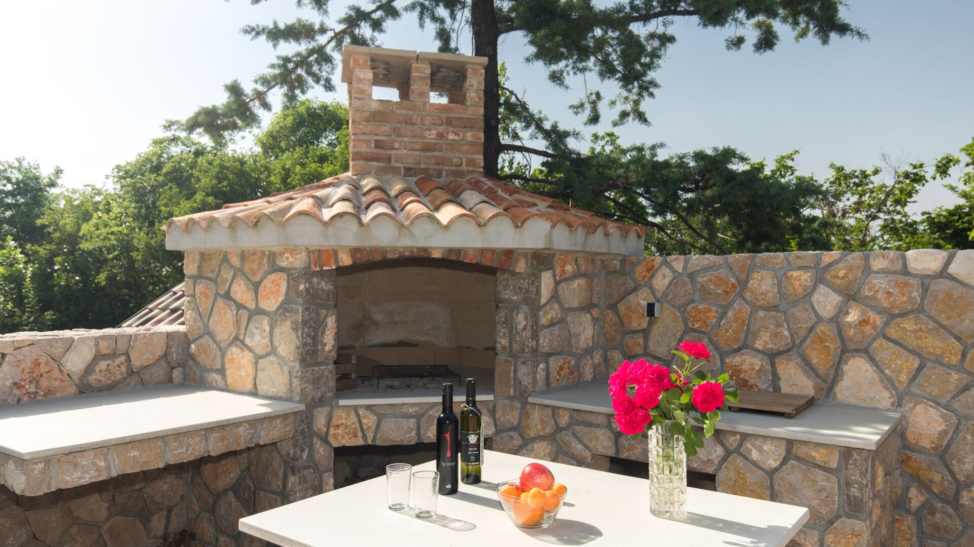 Un four en briques installé à l’extérieur, avec des murs en pierre et un toit recouvert de tuiles, une table ornée d’un vase de fleurs et une bouteille de vin.