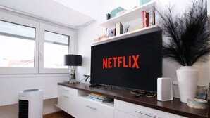A modern living room with a large flat-screen TV displaying "NETFLIX" on the screen, a white air purifier, a lamp, and a bookshelf with various items.