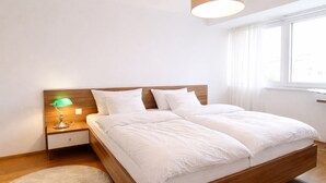 A modern bedroom with a wooden bed frame, white bedding, a green desk lamp, and a round rug.