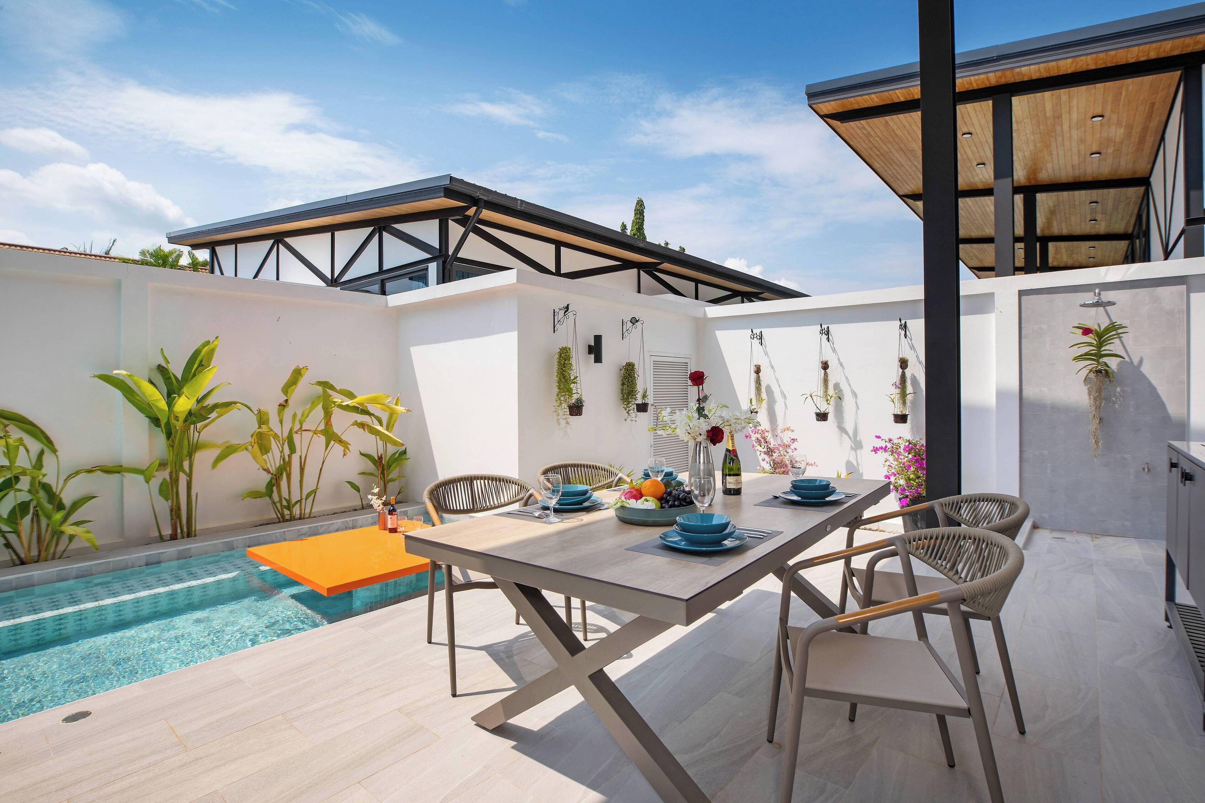 An outdoor dining area with a pool, wooden table, wicker chairs, and potted plants.