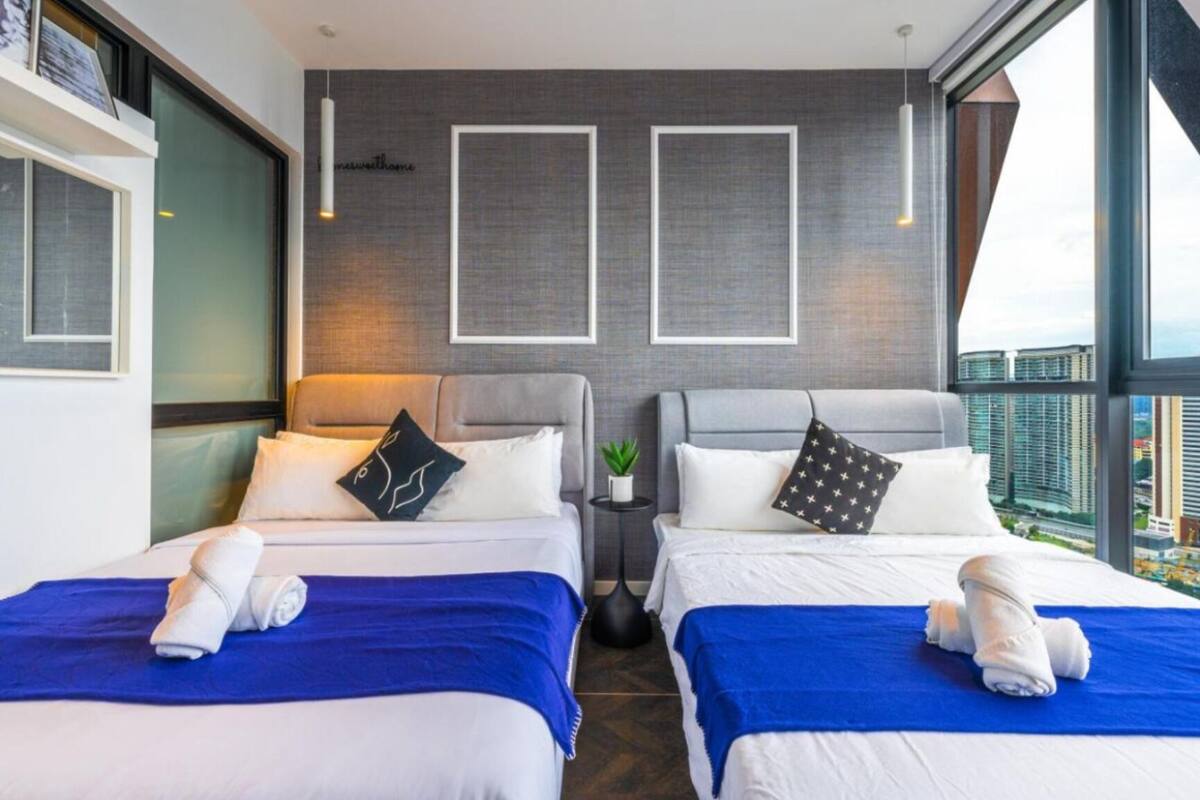 Une chambre d’hôtel moderne avec deux lits, chacun recouvert d’une courtepointe bleue et doté de draps blancs, une petite table de chevet avec une plante, et une vue sur la ville à travers de grandes fenêtres.