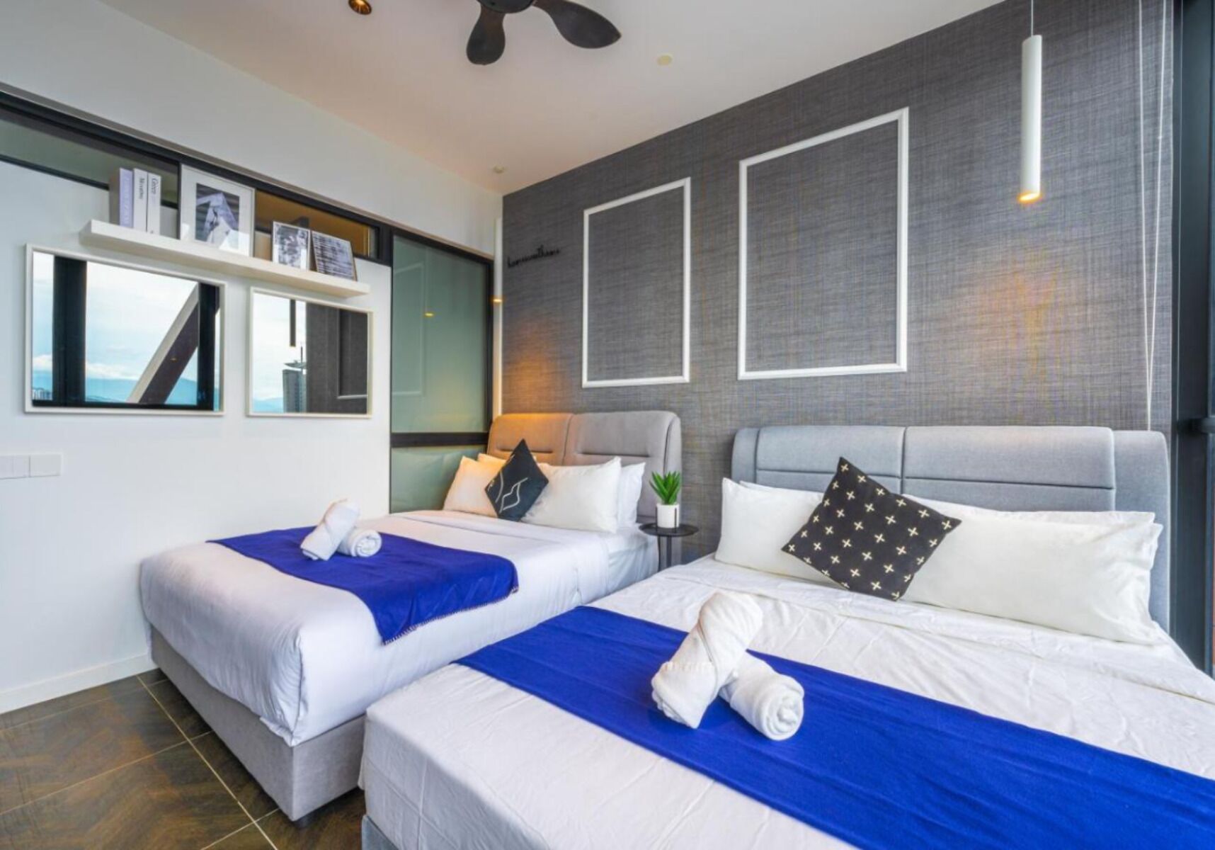 A modern hotel room with two beds, a large window, and a ceiling fan.