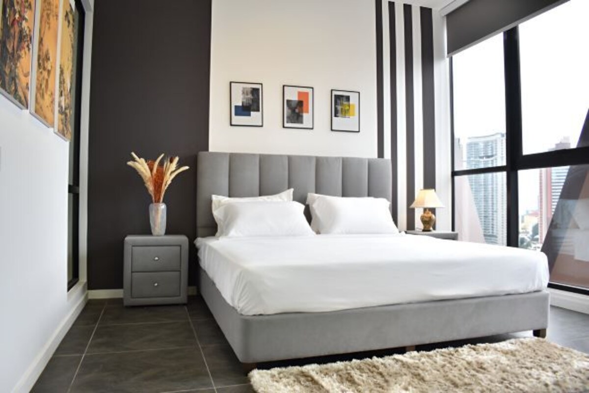 A modern bedroom with a large bed, a nightstand, a vase with dried flowers, and framed pictures on the wall.