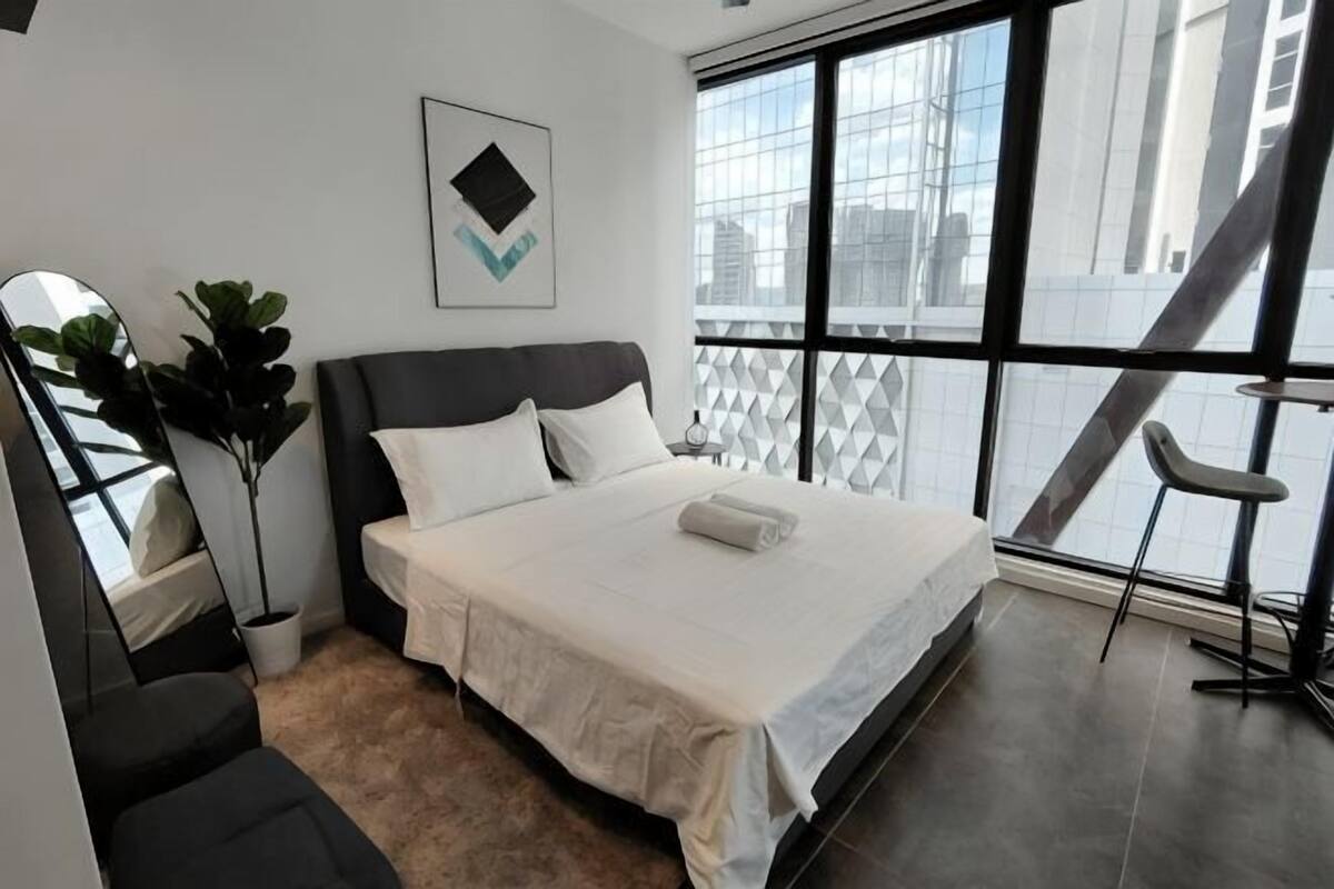 Une chambre moderne avec un grand lit, une plante en pot, un fauteuil et une vue sur la ville grâce à de grandes fenêtres.
