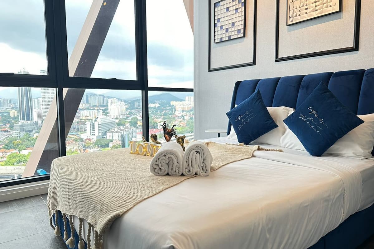Une chambre d’hôtel avec une grande fenêtre, un lit avec du linge de lit blanc, des oreillers bleus et une vue sur le paysage urbain.