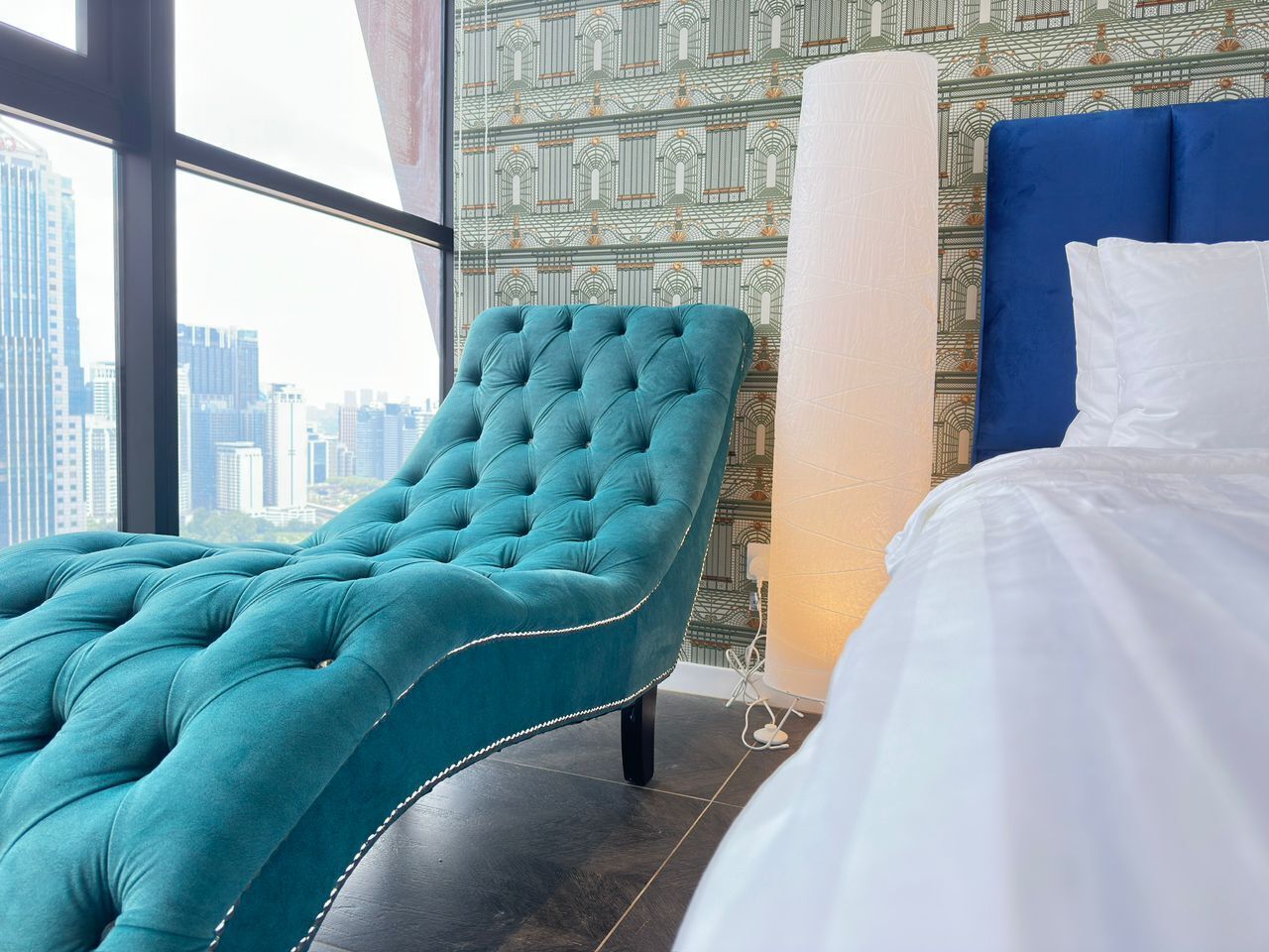 Een moderne hotelkamer met een teal fluweel loungestoel, een groot bed met witte beddengoed en uitzicht op de stad door een groot raam.