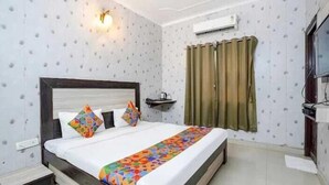 A hotel room with a bed, a nightstand, a wall-mounted shelf, a window with curtains, and an air conditioning unit. - Hotel Taj Royale (Agra)