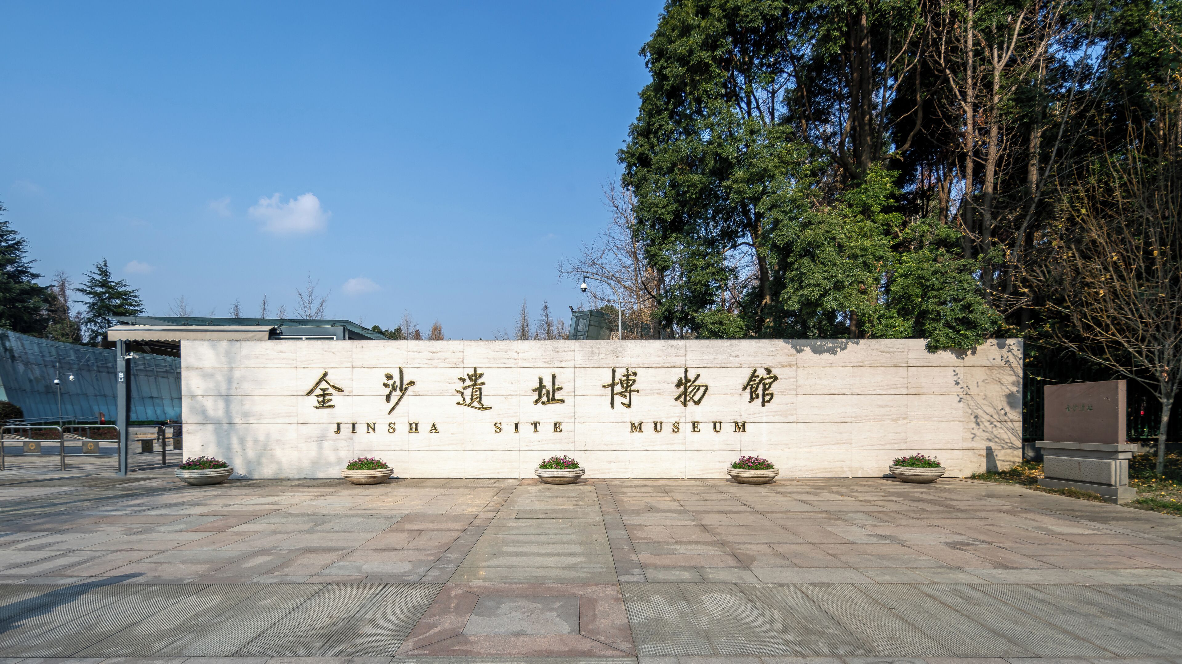 The entrance to Jinsha Site Museum with a stone wall, potted plants, and a clear sky.