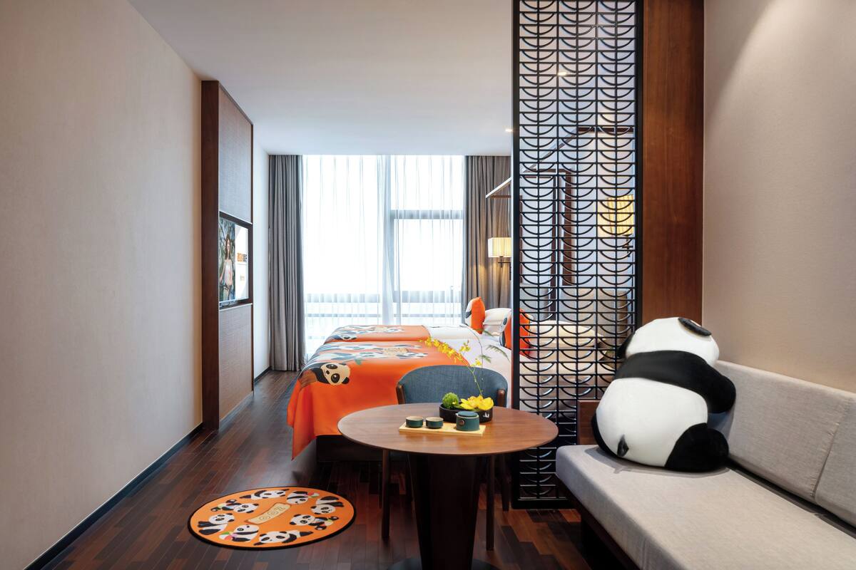 A hotel room with a bed, sofa, table, and decorative panda plush toy.