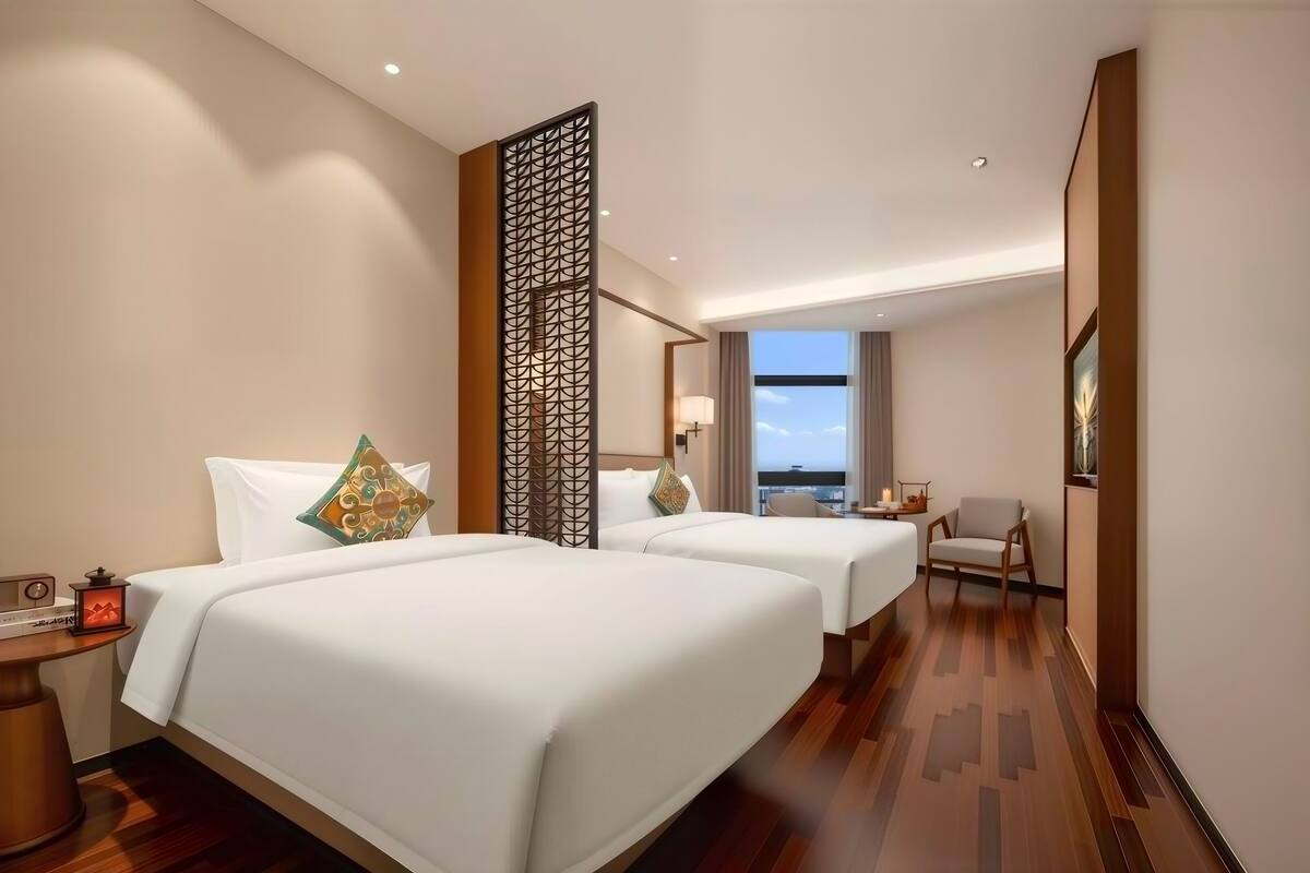 A hotel room with two beds, a nightstand, a chair, a decorative screen, and a view of the sky.