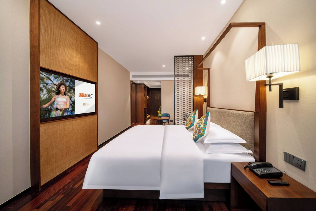 A hotel room with a large bed, a bedside table with a lamp, a phone, and a wall-mounted TV.