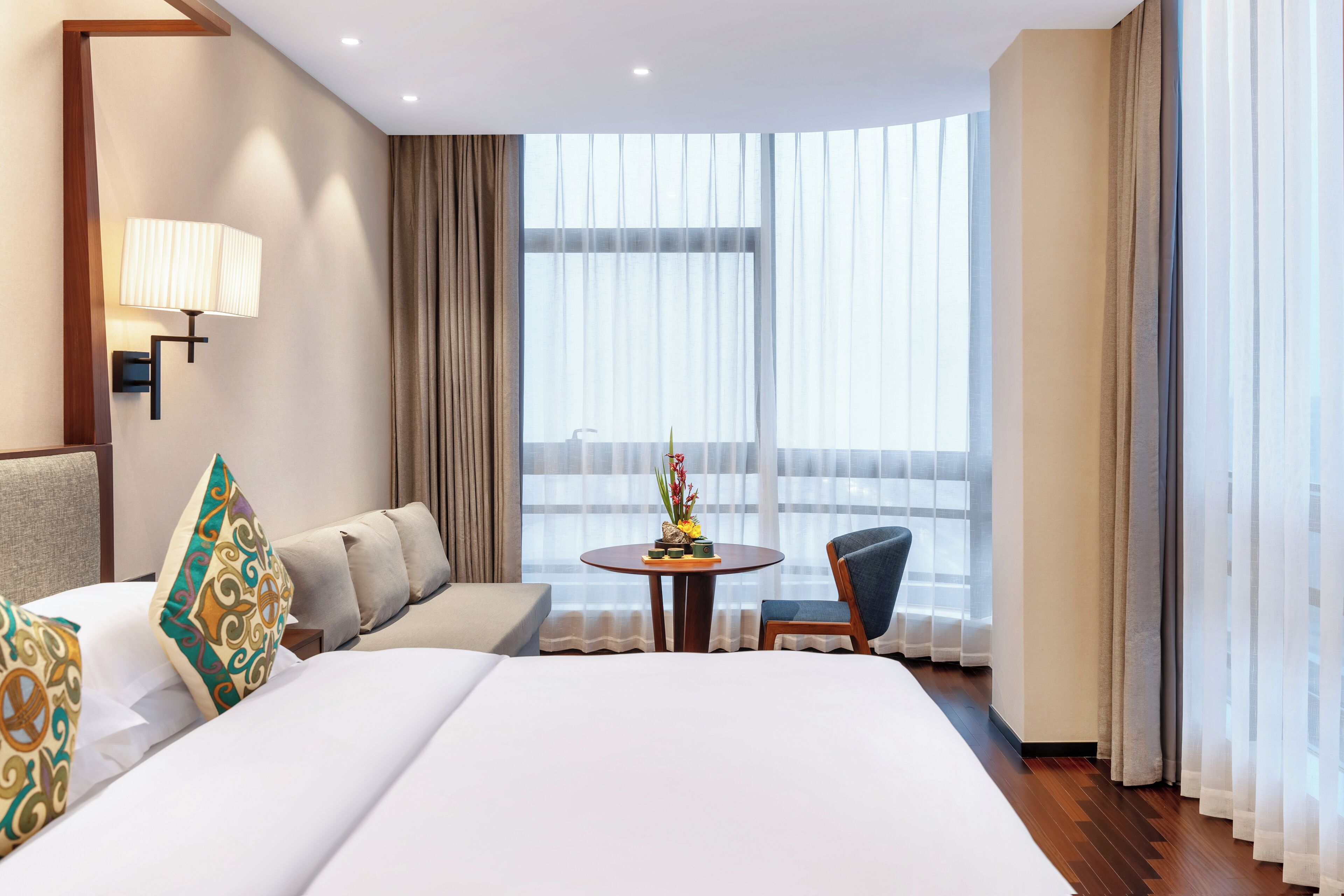 A hotel room with a large bed, a sofa, a small table with a vase, and a chair.