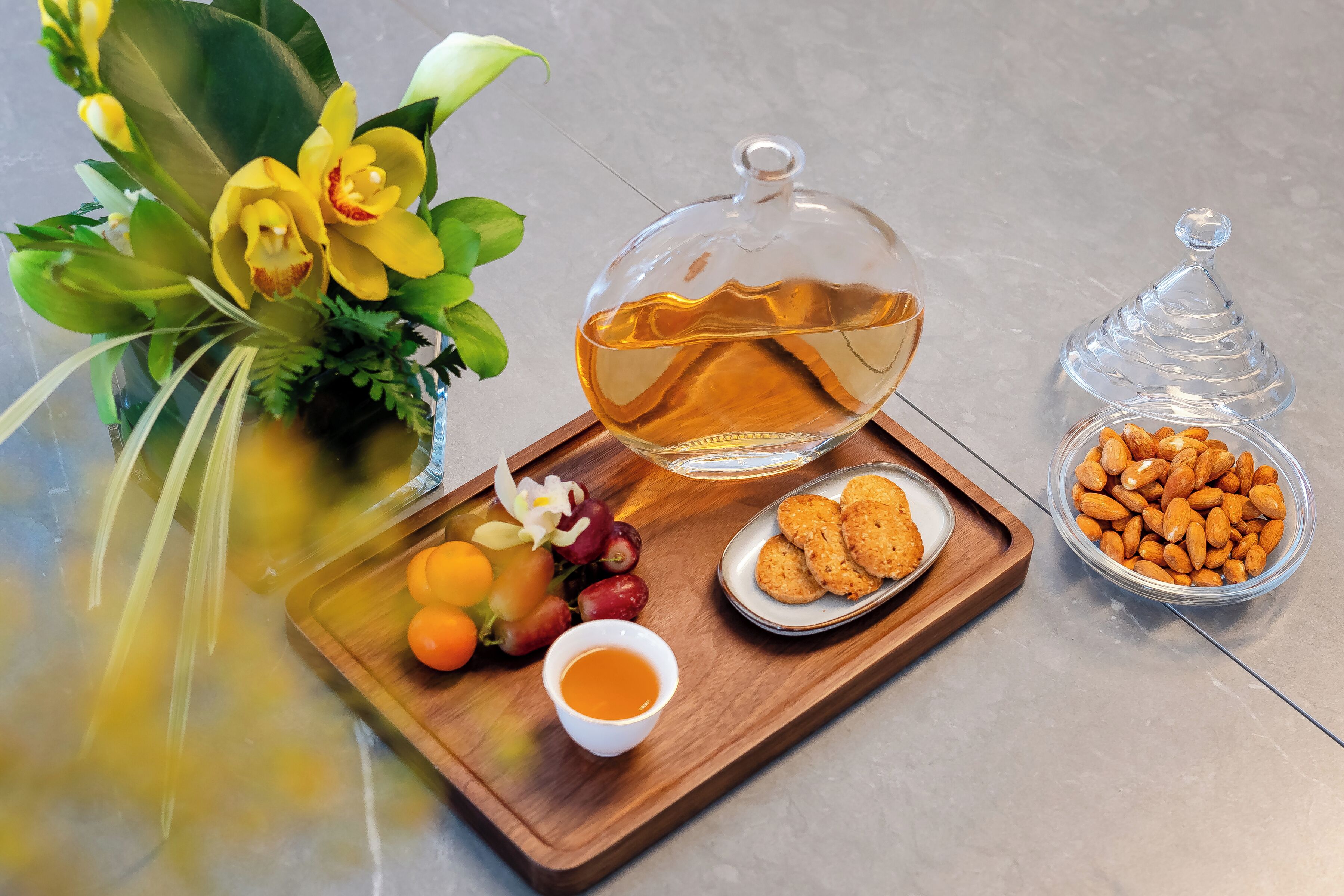 A wooden tray with a glass bowl of honey, a small bowl of almonds, a plate of cookies, and a vase with flowers.