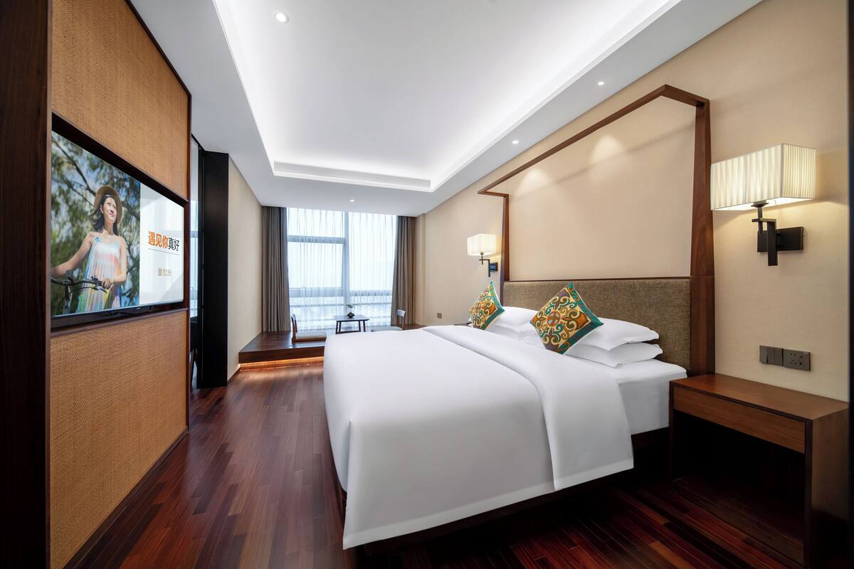 A modern hotel room with a large bed, a TV mounted on the wall, a bedside table with a lamp, and a view of the outdoors through the window.