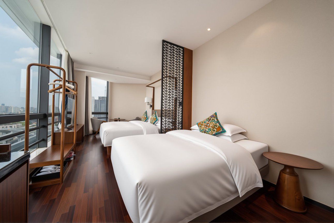 Een hotelkamer met twee bedden, een houten vloer, een groot raam met uitzicht op de stad en een decoratieve roomdivider.