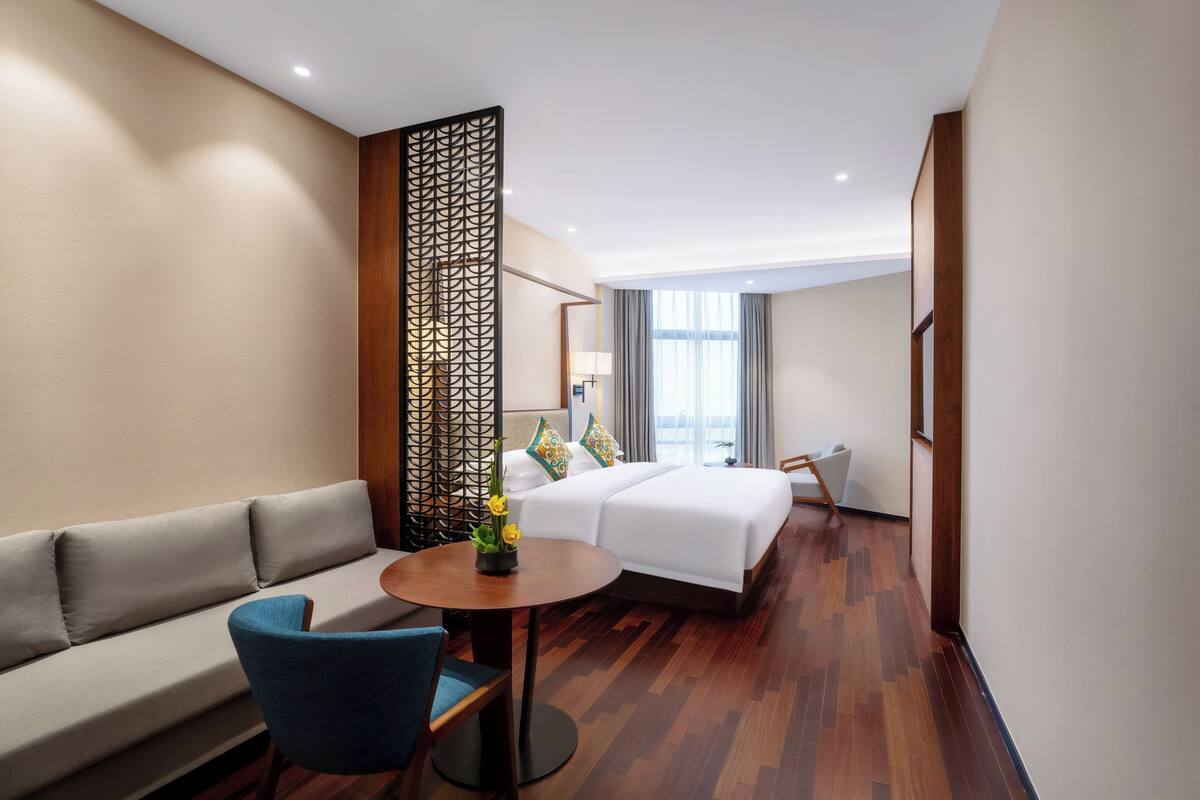 A hotel room with a sofa, a bed with pillows, a desk with a chair, and a decorative partition.