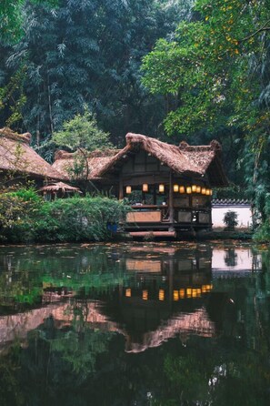 A traditional wooden structure with thatched roofs, surrounded by lush greenery and a calm water body reflecting the building. - SSAW Boutique Hotel Swufe Chengdu (Chengdu)