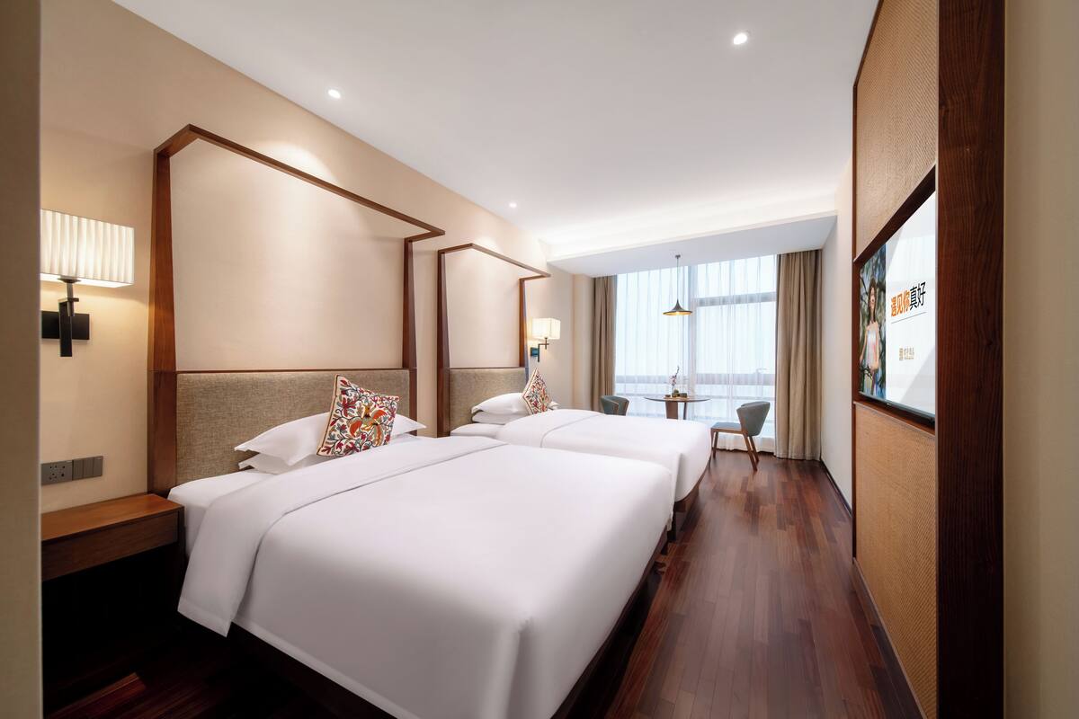 A hotel room with two beds, a desk, a chair, and a large window with curtains.