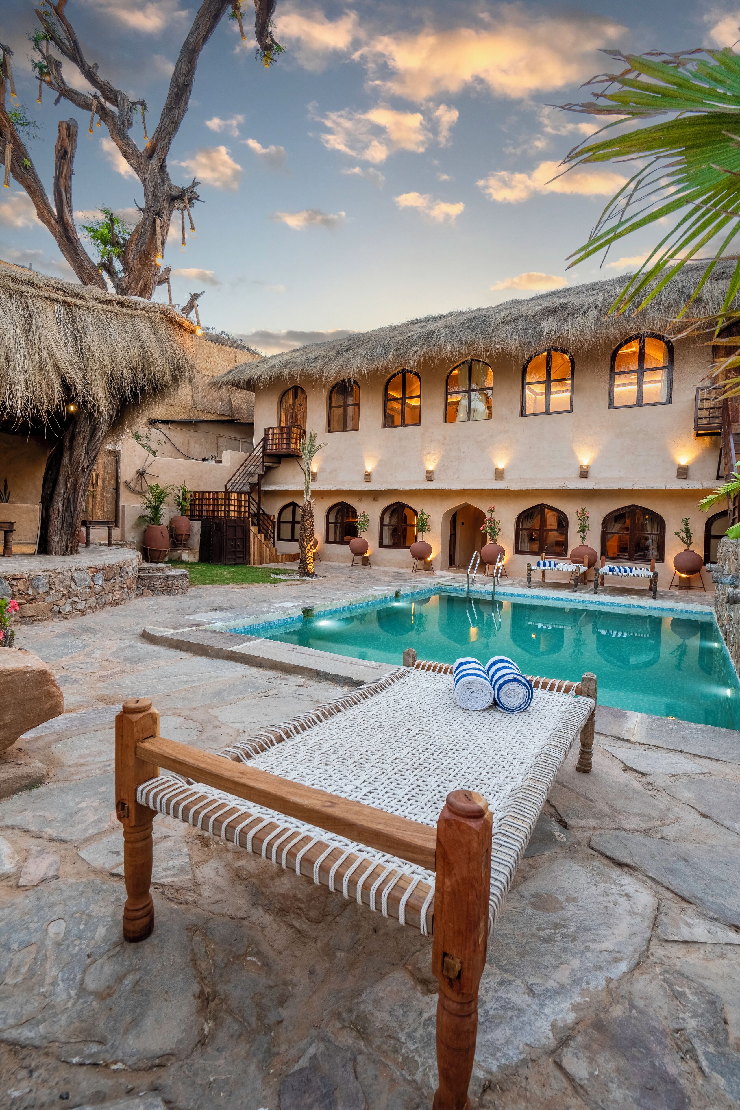 A pool area with a wooden bench and lounge chairs, surrounded by a building with arched windows and a thatched roof.