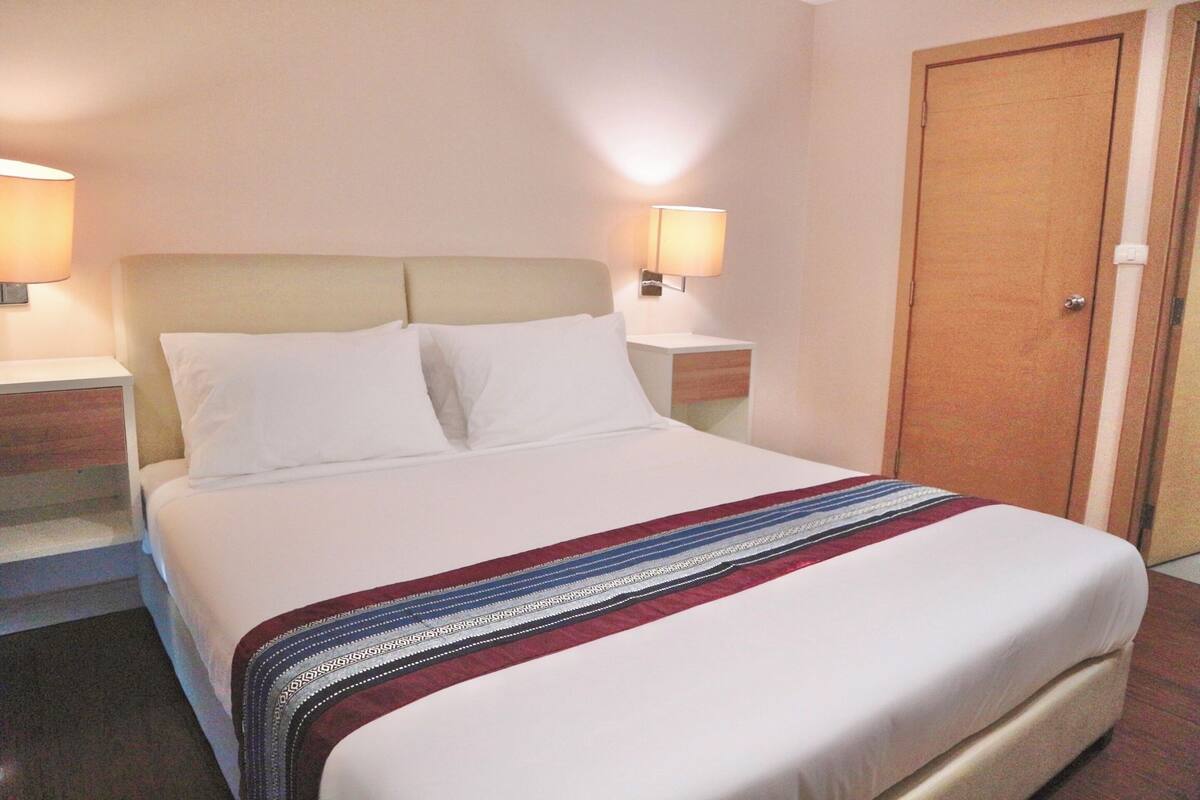 Een net opgemaakt bed met witte lakens en een decoratieve deken, twee bedlampen en een houten deur.