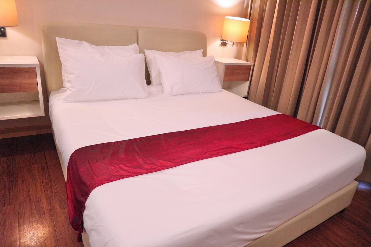 Een net opgemaakt bed met witte lakens en een rode accentkleur, met aan weerszijden houten nachtkastjes met lampen, in een kamer met houten vloer en gordijnen.