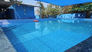 A clear swimming pool with a tiled edge, surrounded by a shaded area and a building with a blue facade. - Hotel Hari Om Swimming Pool In Diu (Diu)
