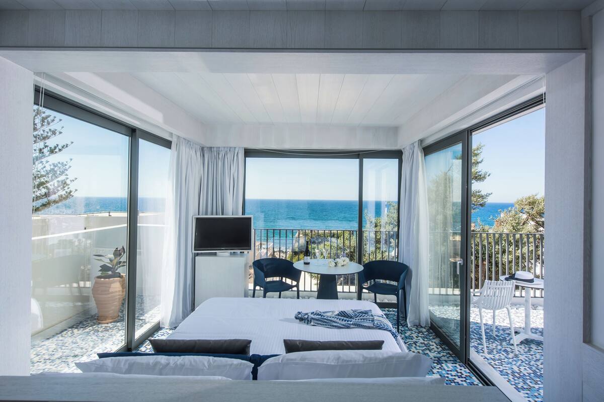 A hotel room with a bed, a TV, a balcony with a table and chairs, and a view of the sea.
