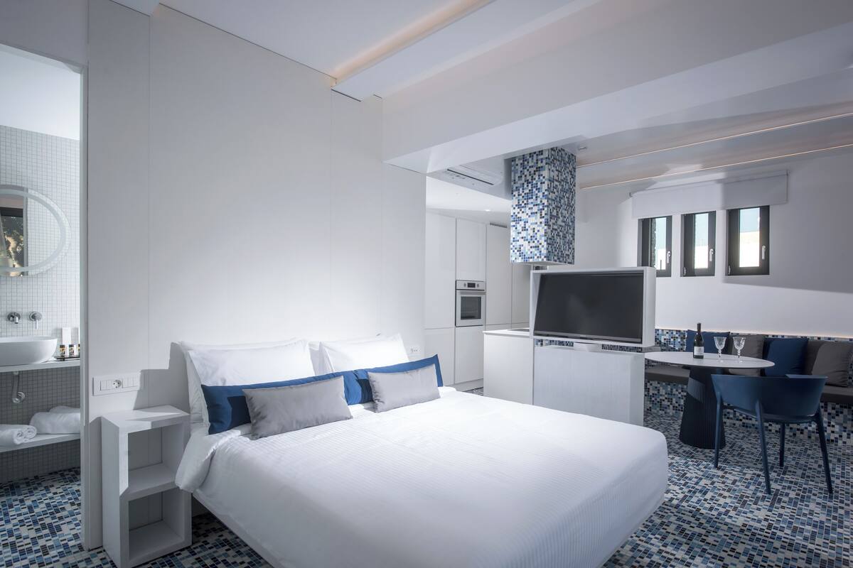 A modern hotel room with a large bed, a TV, a desk with a chair, a bathroom with a sink and mirror, and a patterned floor.