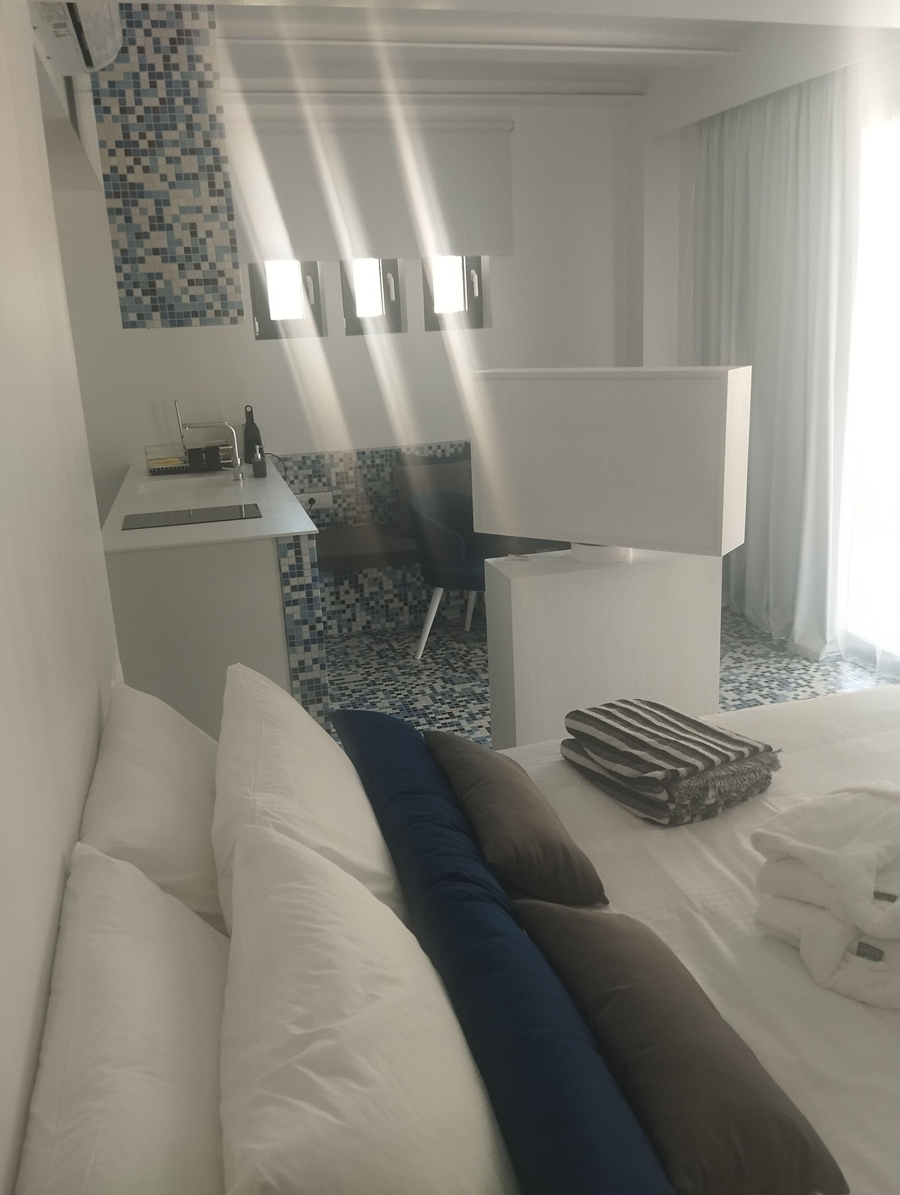 Ein Hotelzimmer mit Bett, Kissen, einem kleinen Schreibtisch mit Waschbecken und einem durch eine Glastrennwand abgetrennten Badezimmer.