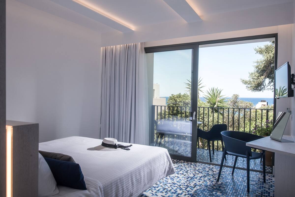 A hotel room with a bed, a desk, and a balcony with a view of the sea.