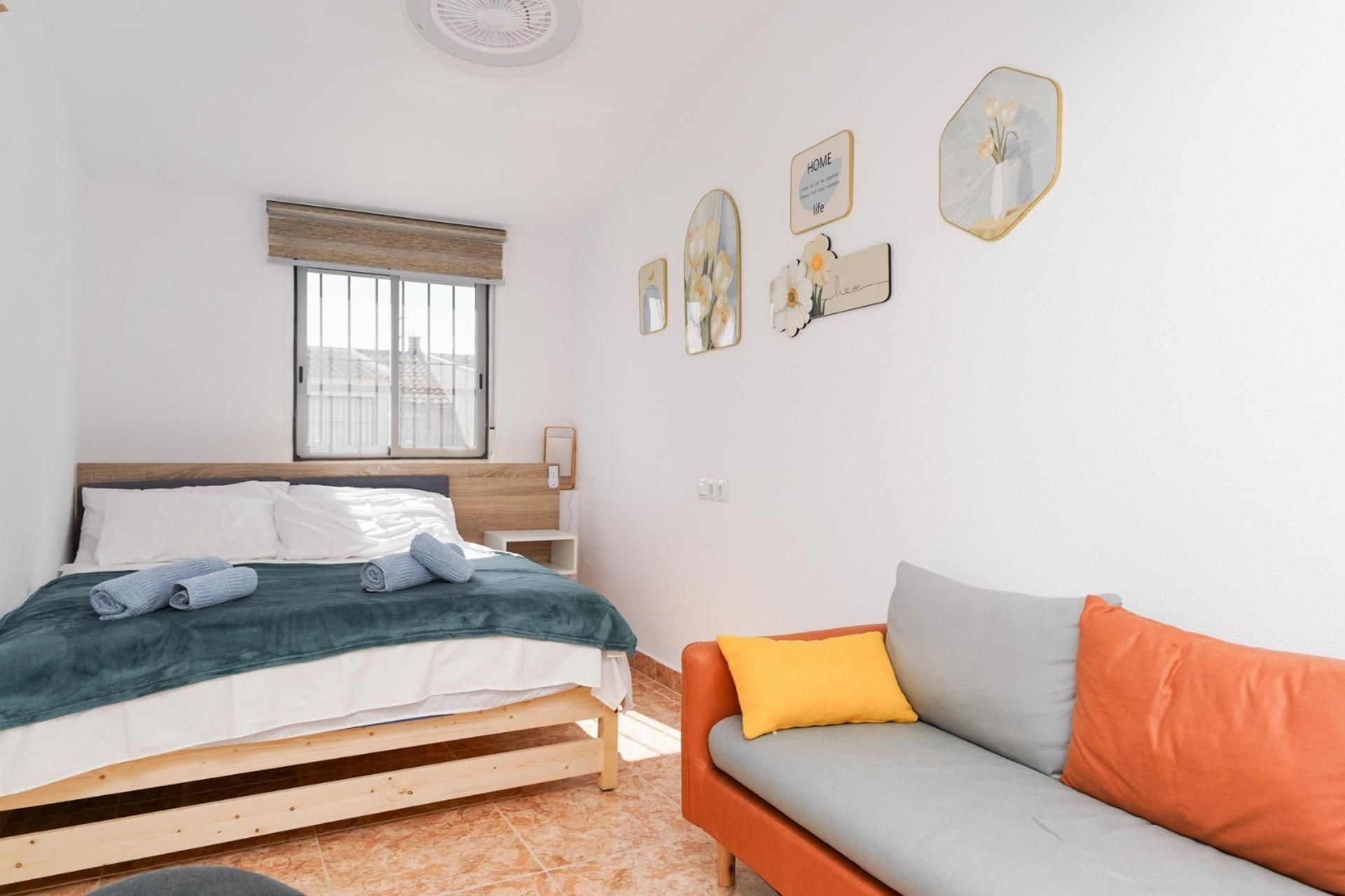 A bedroom with a bed, a window, a bedside table, a nightstand, a sofa, and wall decorations.
