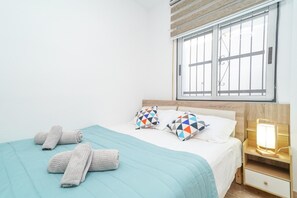 Una cama bien hecha con una colcha azul, dos almohadas estampadas y dos toallas enrolladas.