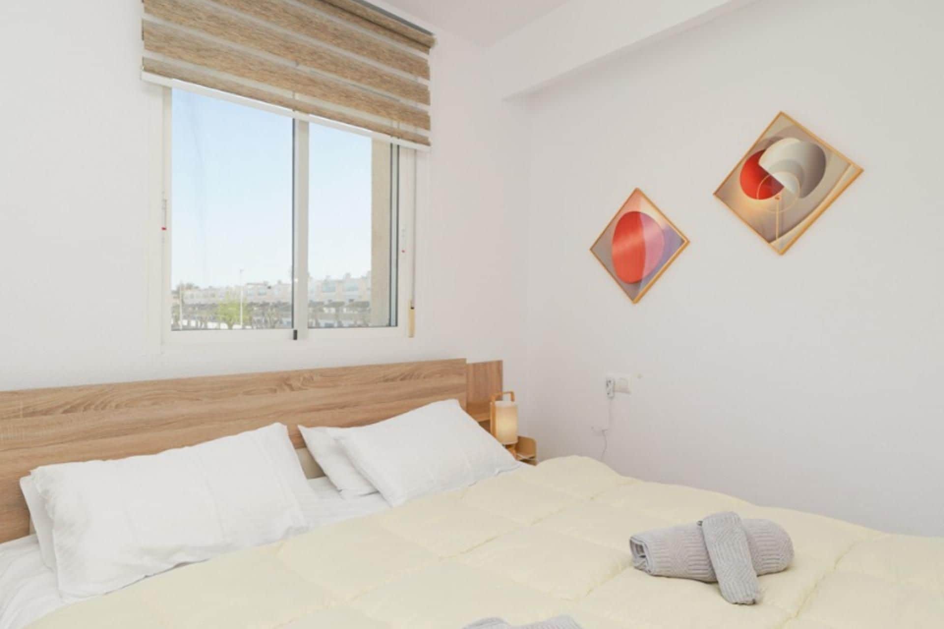 Een slaapkamer met een groot bed, twee kussens, een nachtkastje met een lamp en een raam met uitzicht op gebouwen.