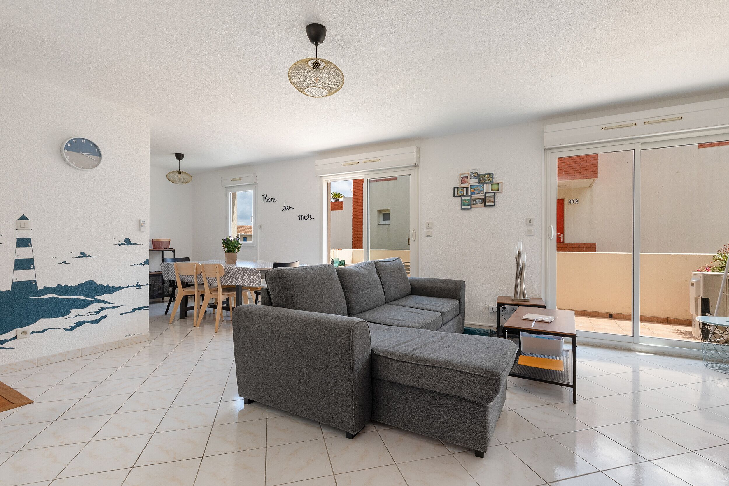 A modern living room with a grey sofa, a dining area with a table and chairs, and a large sliding glass door leading to an outdoor area.