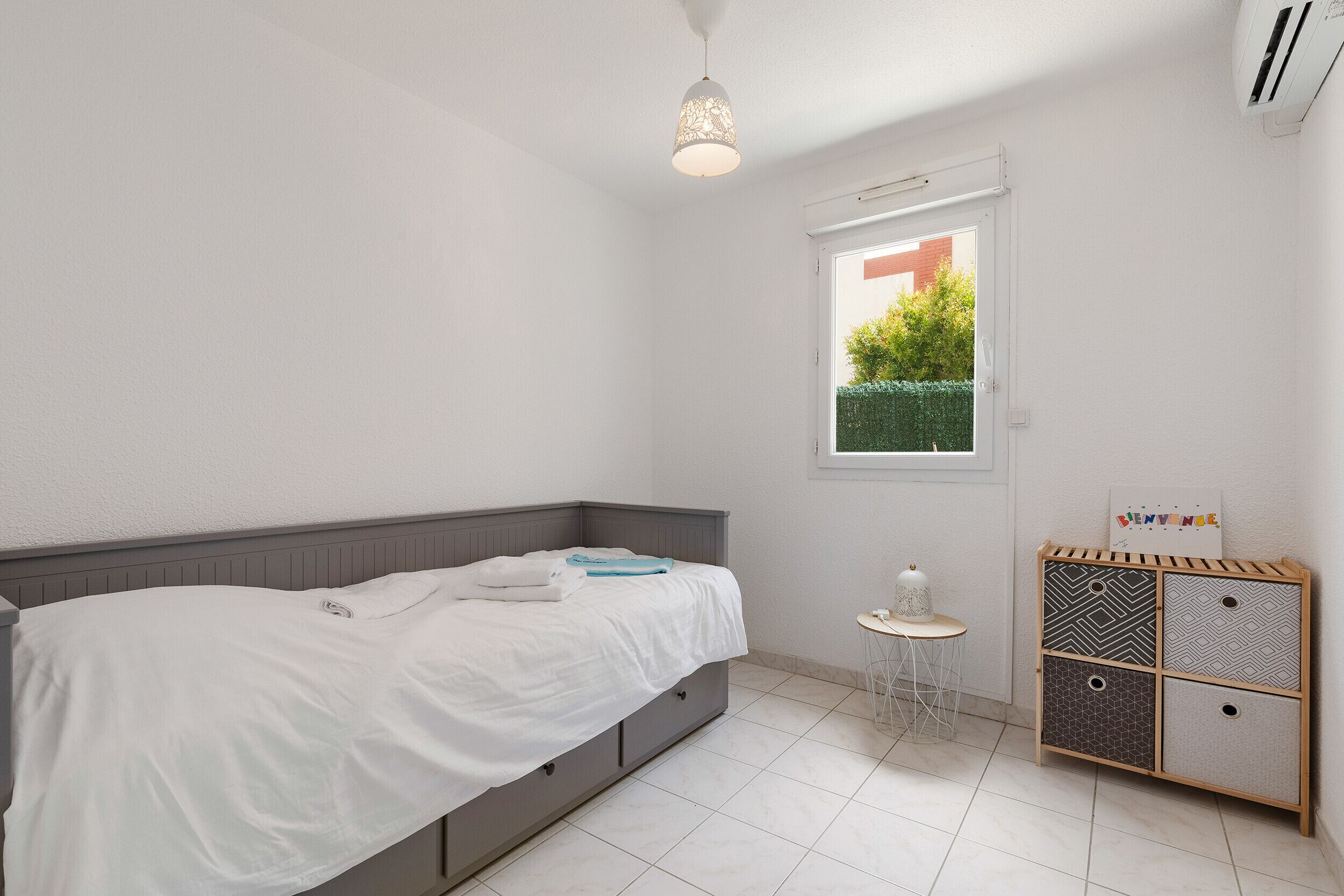 Een slaapkamer met een bed, een raam, een klein tafeltje en een opbergmeubel.