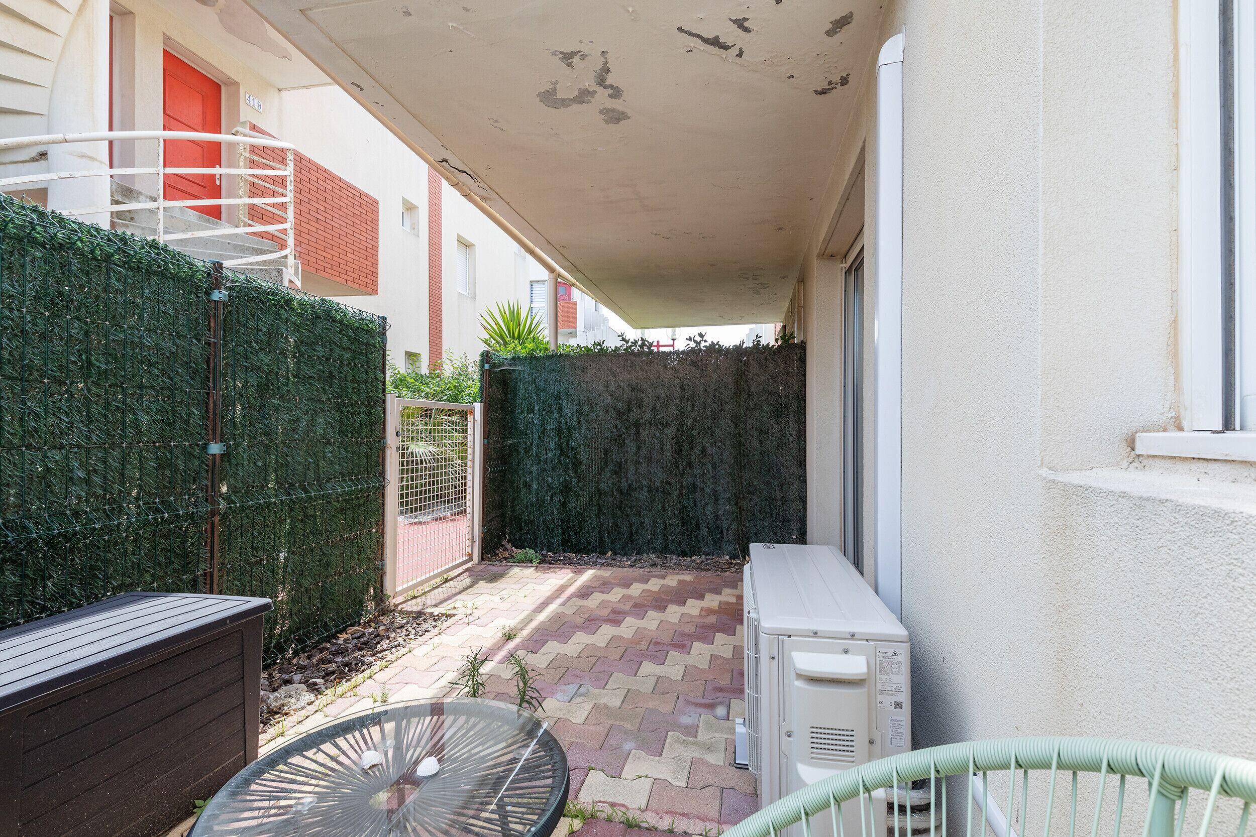 Un estrecho pasillo exterior con suelo de baldosas, un seto verde, una pared blanca y una puerta roja.