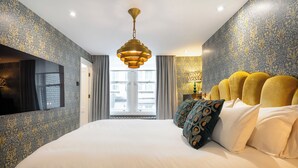 A hotel room with a large bed, a modern chandelier, patterned wallpaper, and a view of the city through the window.