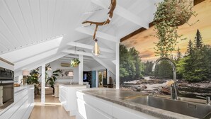 Moderni keittiö, jossa on suuri keittiösaareke, riippuvalaisin ja metsämaiseman kuvaava taulu.