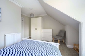 A bedroom with a sloped ceiling, a bed with a striped bedspread, a white wardrobe, a grey chair, and a small wooden shelf. - Lade Braes No 35 | St Andrews (St Andrews)