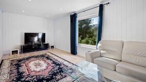 A modern living room with a large flat-screen TV, a glass coffee table, a beige sofa, and a patterned rug. - Entire Single-family Home - Centrally Located in Voss (Voss)