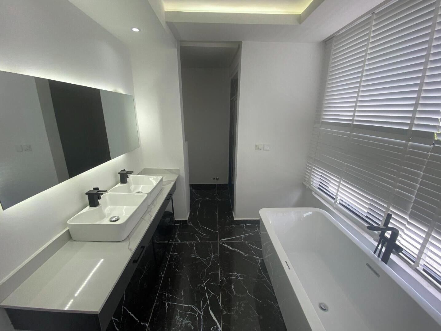 A modern bathroom with a large mirror, two sinks, a bathtub, and a window with blinds.