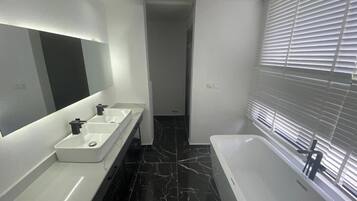 A modern bathroom with a large mirror, two sinks, a bathtub, and a window with blinds.